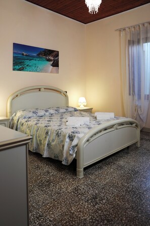 A bedroom with a bed, a nightstand, a lamp, a painting of a beach scene, and a window with curtains. - Annalú Vacation Home (I.U.N. R5074) (Orosei)
