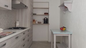 A narrow kitchen with light wood cabinets, a gas stove, a coffee maker, and a small table with fruit. - Annalú Holiday House (I.U.N. R5074) (Orosei)