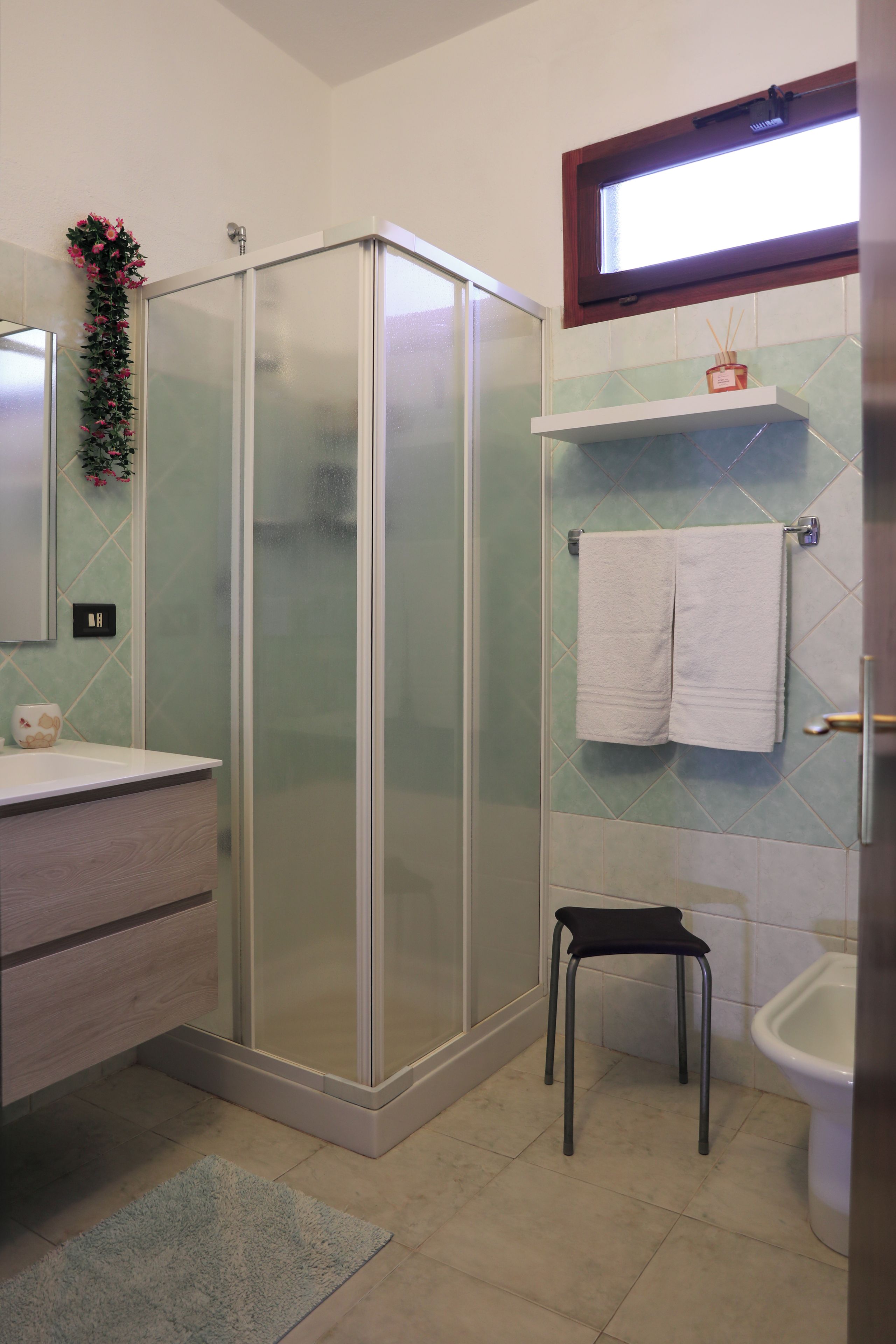 A bathroom with a glass-enclosed shower, a sink with a mirror, a toilet, and a small stool.