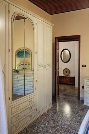 A room with a large arched mirror, a wardrobe with multiple drawers, a small round mirror on the wall, and a tiled floor. - Annalú Vacation Home (I.U.N. R5074) (Orosei)