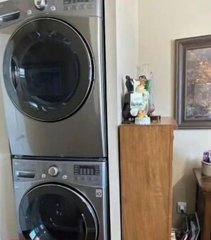 Une machine à laver et un sèche-linge superposés, placés dans une pièce où se trouve un meuble en bois et une petite statue posée sur le dessus.