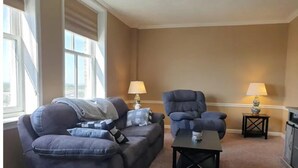 A living room with a dark blue sofa, a matching armchair, and a small black coffee table. There are two lamps on side tables, and the room has large windows with blinds.
