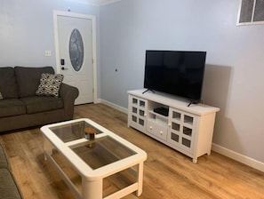 Una sala de estar con un sofá, una mesa de centro de vidrio, un mueble blanco para televisor y una puerta con panel de vidrio decorativo.