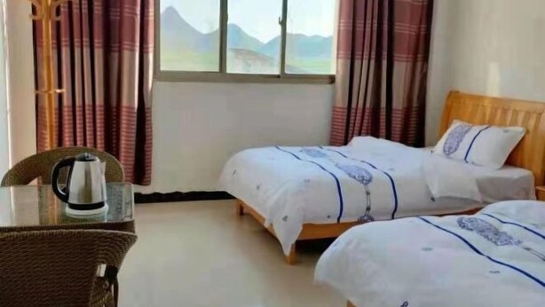 A room with two beds, a wooden headboard, a glass table with a kettle, and a view of mountains through the window. - Jinfeng Farmhouse (Luoping)