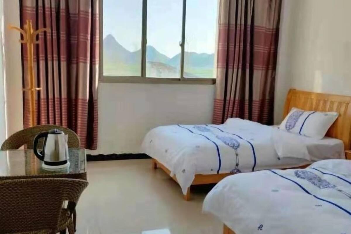 A room with two beds, a wooden headboard, a glass table with a kettle, and a view of mountains through the window.