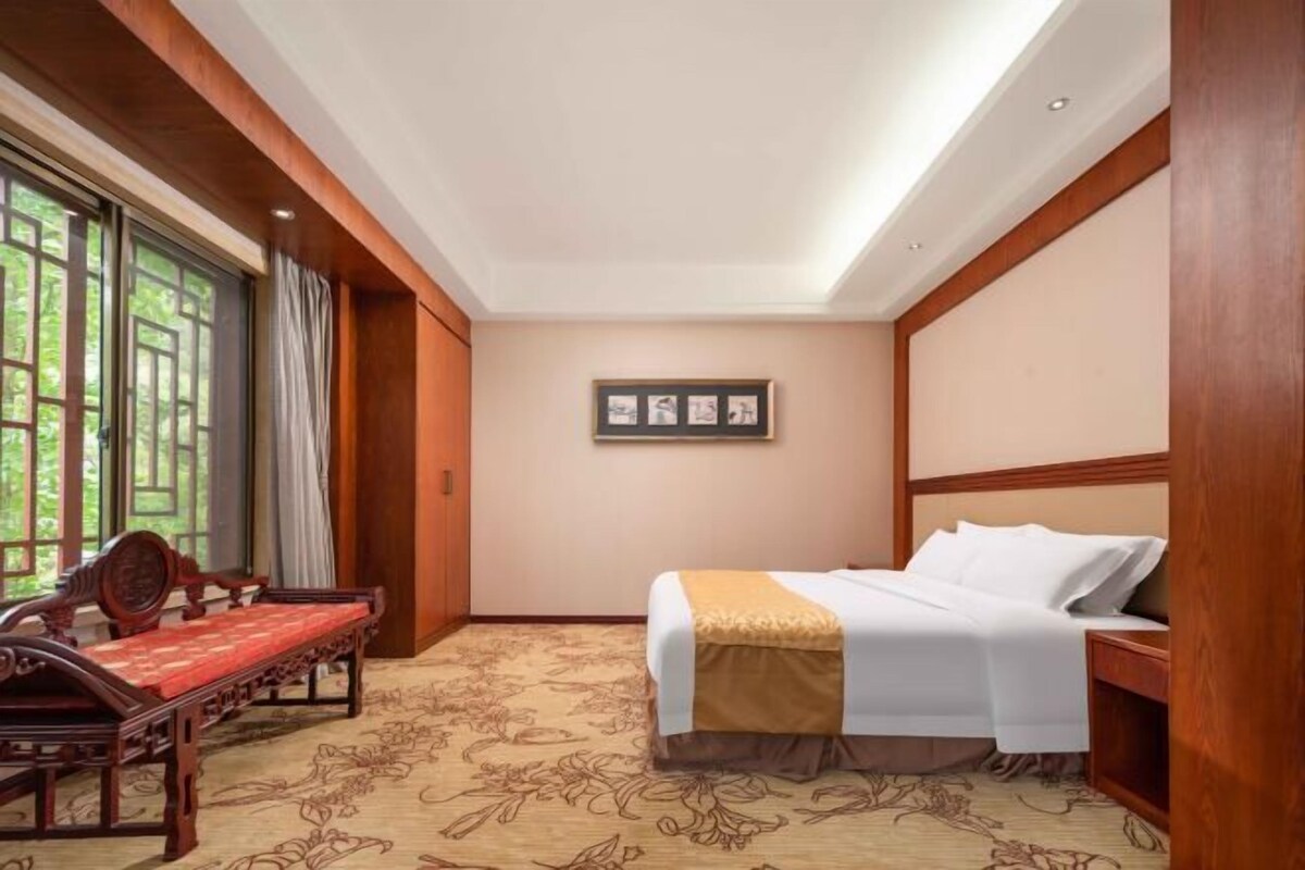 Habitación de hotel con cama, un banco de madera, un cuadro en la pared y una ventana con cortinas.