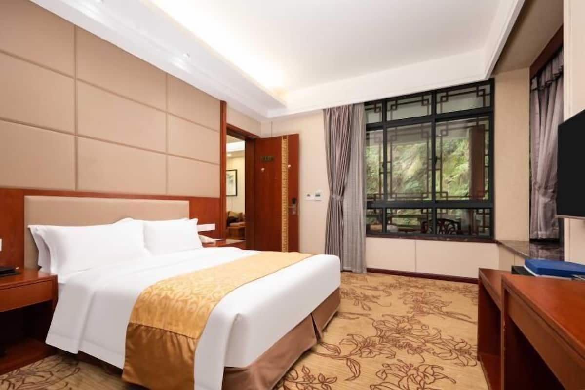 A hotel room with a large bed, a wooden headboard, a TV, a desk, and a window with curtains.