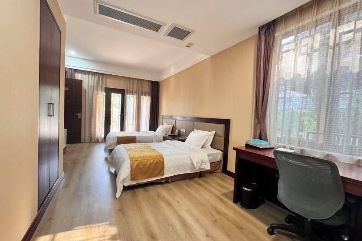 A hotel room with two beds, a desk, a chair, and a large window with curtains.