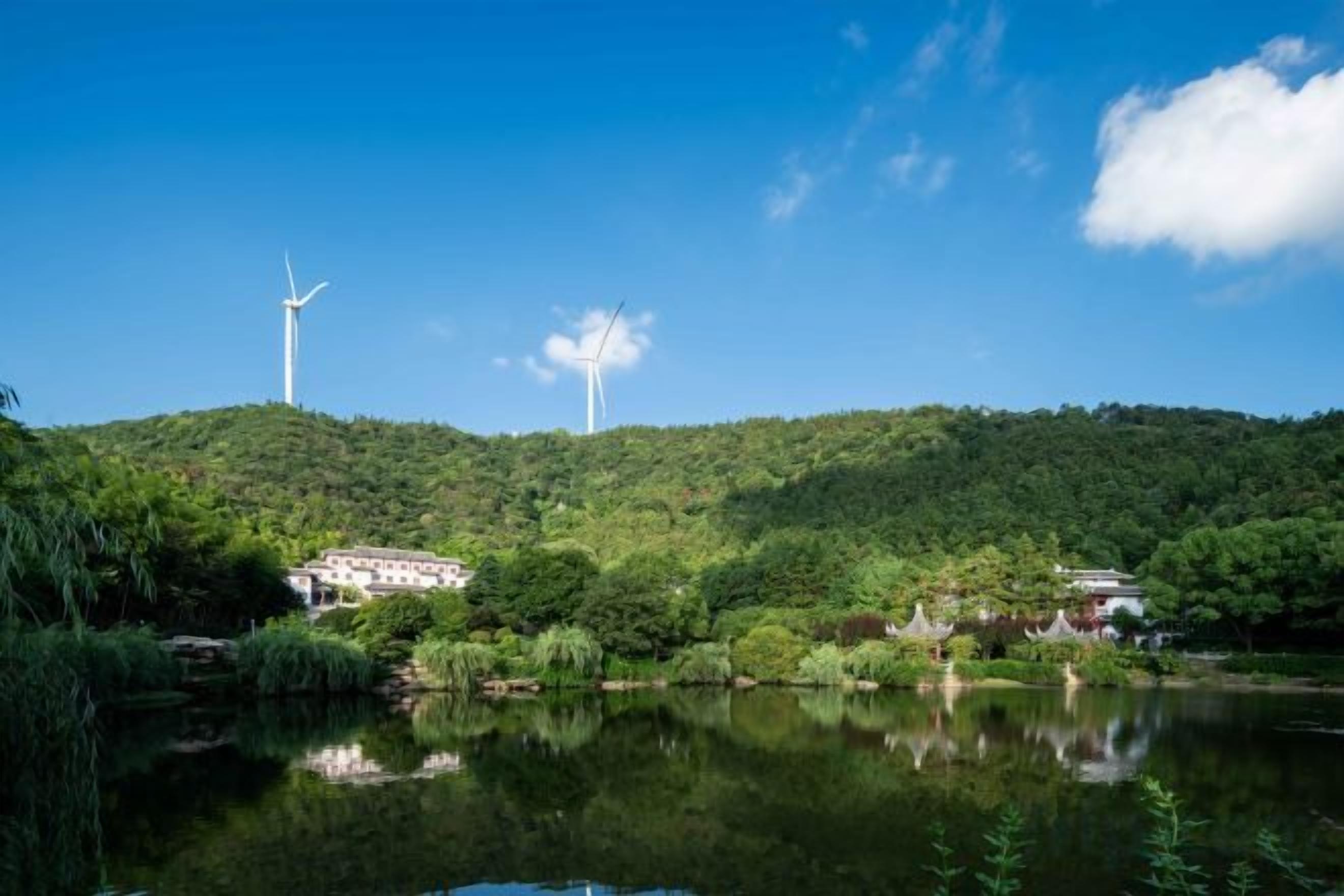 Un paisaje tranquilo con un lago, dos aerogeneradores y edificios tradicionales en una colina.