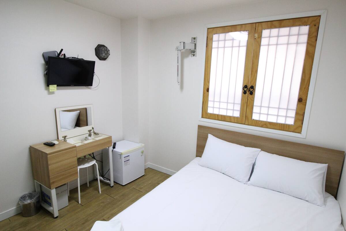 Habitación de hotel con cama, escritorio con espejo, refrigerador pequeño y ventana con contraventanas de madera.