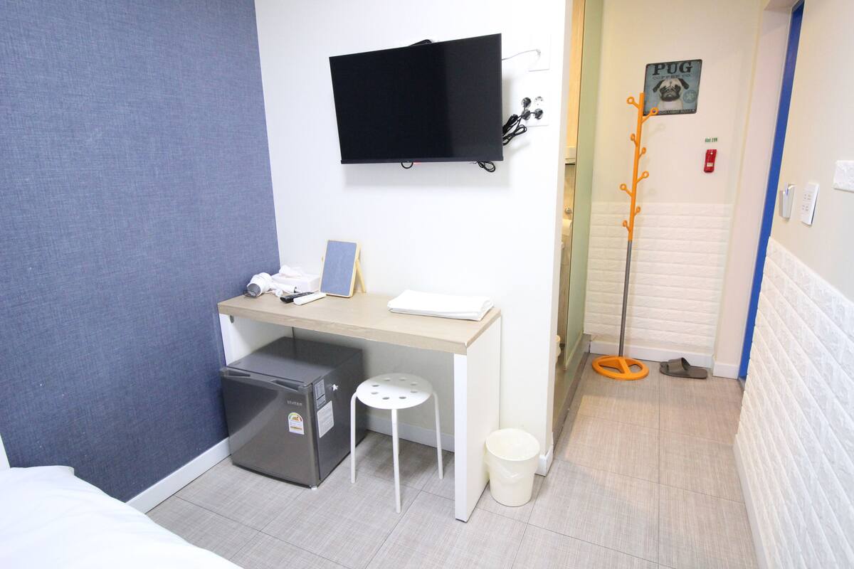 Habitación de hotel compacta con cama, escritorio con computadora, refrigerador pequeño, basurero blanco y televisor montado en la pared.