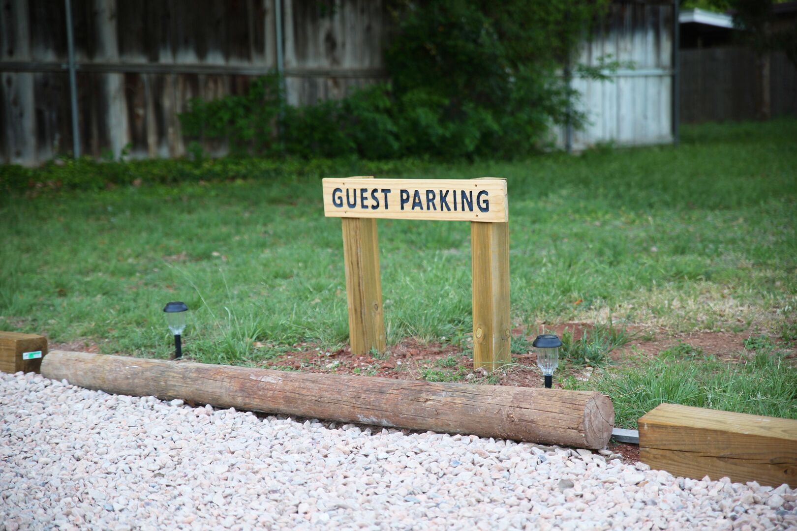A wooden sign labeled "GUEST PARKING" in a grassy area with gravel and wooden logs.
