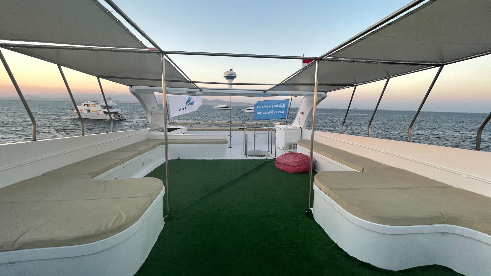 A boat deck with a green carpet, cushioned seating, and a banner advertising a website.