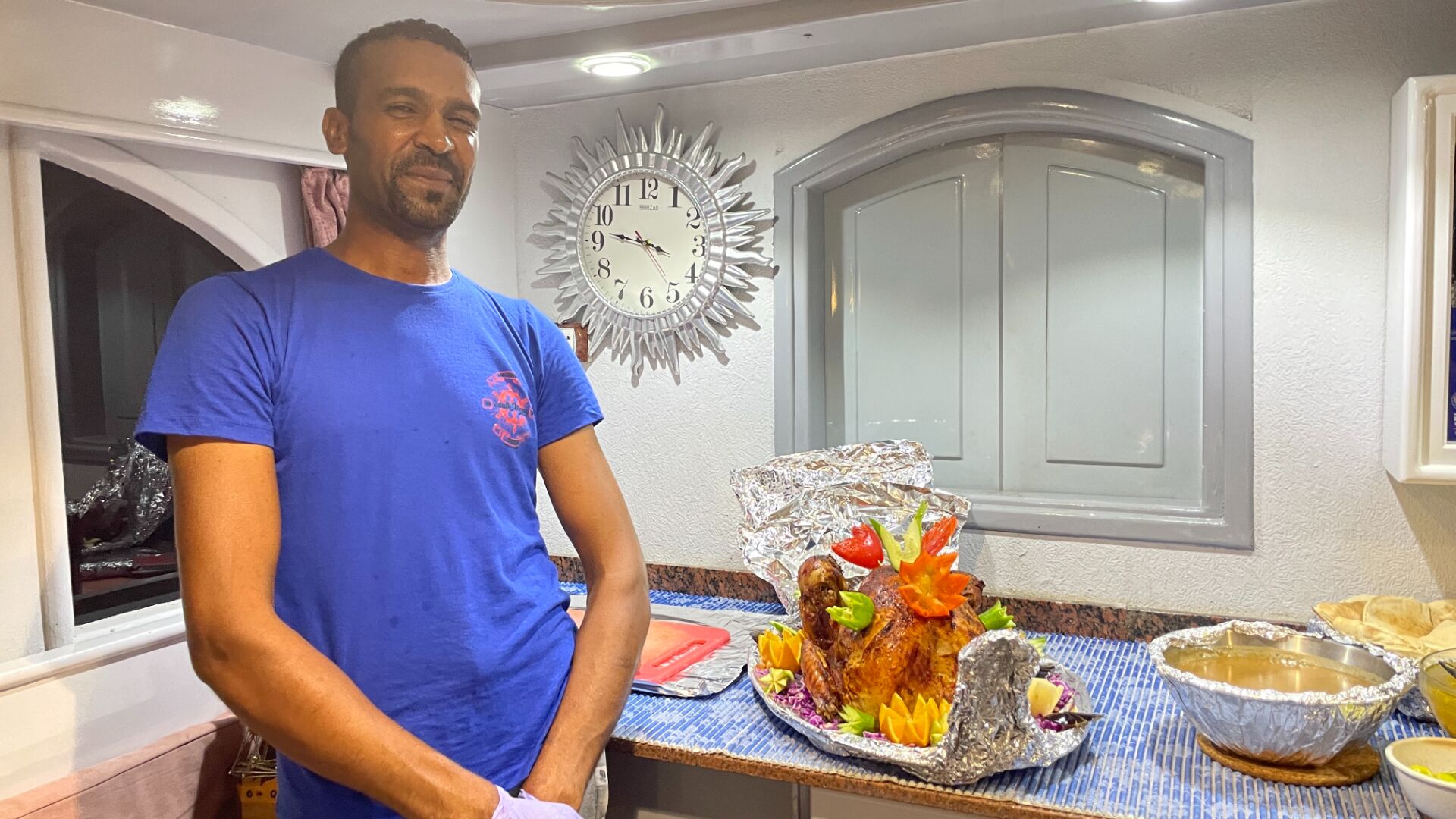 Un homme vêtu d’une chemise bleue se tient dans une cuisine. On peut y voir une grande horloge décorative accrochée au mur, une dinde rôtie posée sur le comptoir et un bol de soupe.
