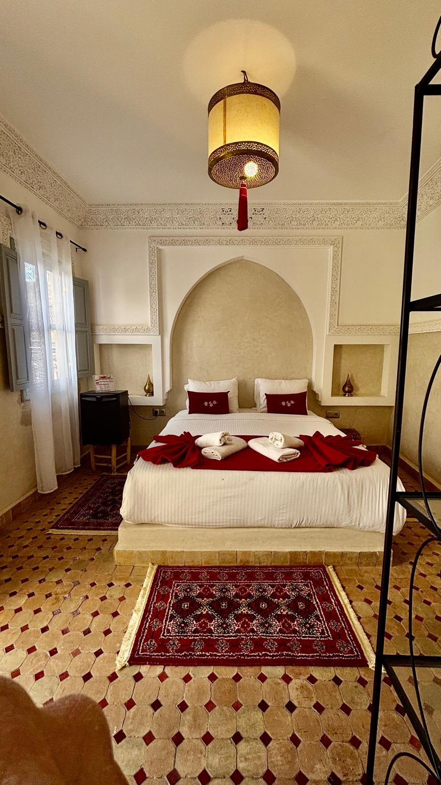 A hotel room with a large bed, a chandelier, a red and white patterned rug, and a decorative ceiling.