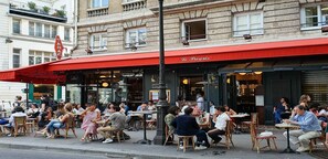 Un café en plein air avec des tables et des chaises, un auvent rouge et une enseigne indiquant « Le Progrès ».