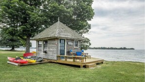 A small wooden cabin with a deck, kayaks, and a chair by the water. - Relaxing Lake Waterfront Home (Simcoe Island, Frontenac Islands)