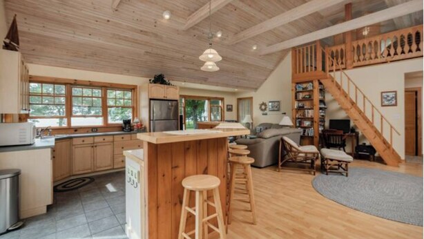 A spacious kitchen with wooden cabinets, a central island with stools, a large refrigerator, and a dining area with a high wooden staircase. - Relaxing Lake Waterfront Home (Simcoe Island, Frontenac Islands)