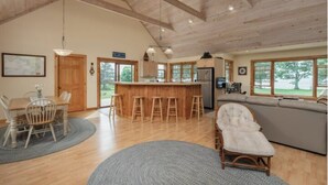 A spacious living area with a wooden ceiling, a dining table with chairs, a bar with stools, a large window, and a sofa. - Relaxing Lake Waterfront Home (Simcoe Island, Frontenac Islands)