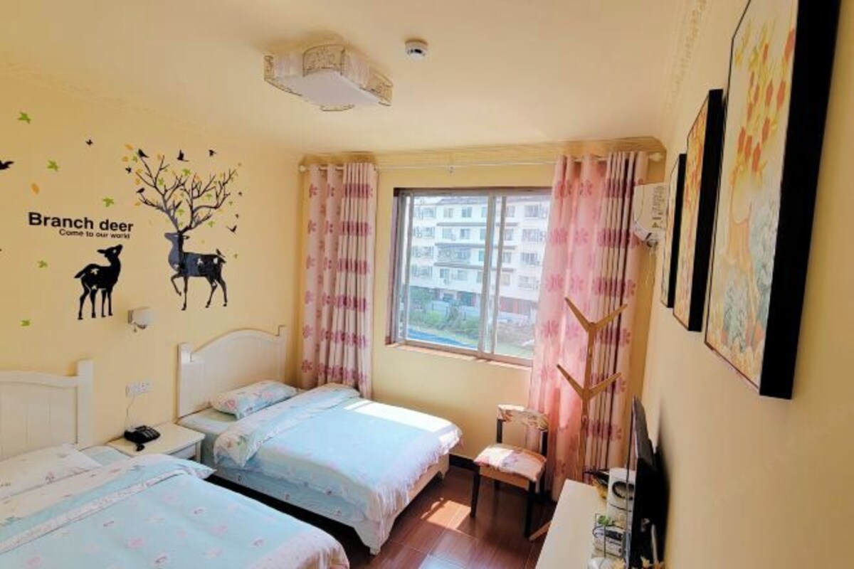 A room with two beds, a chair, a window with curtains, and a wall decal of a tree and deer.