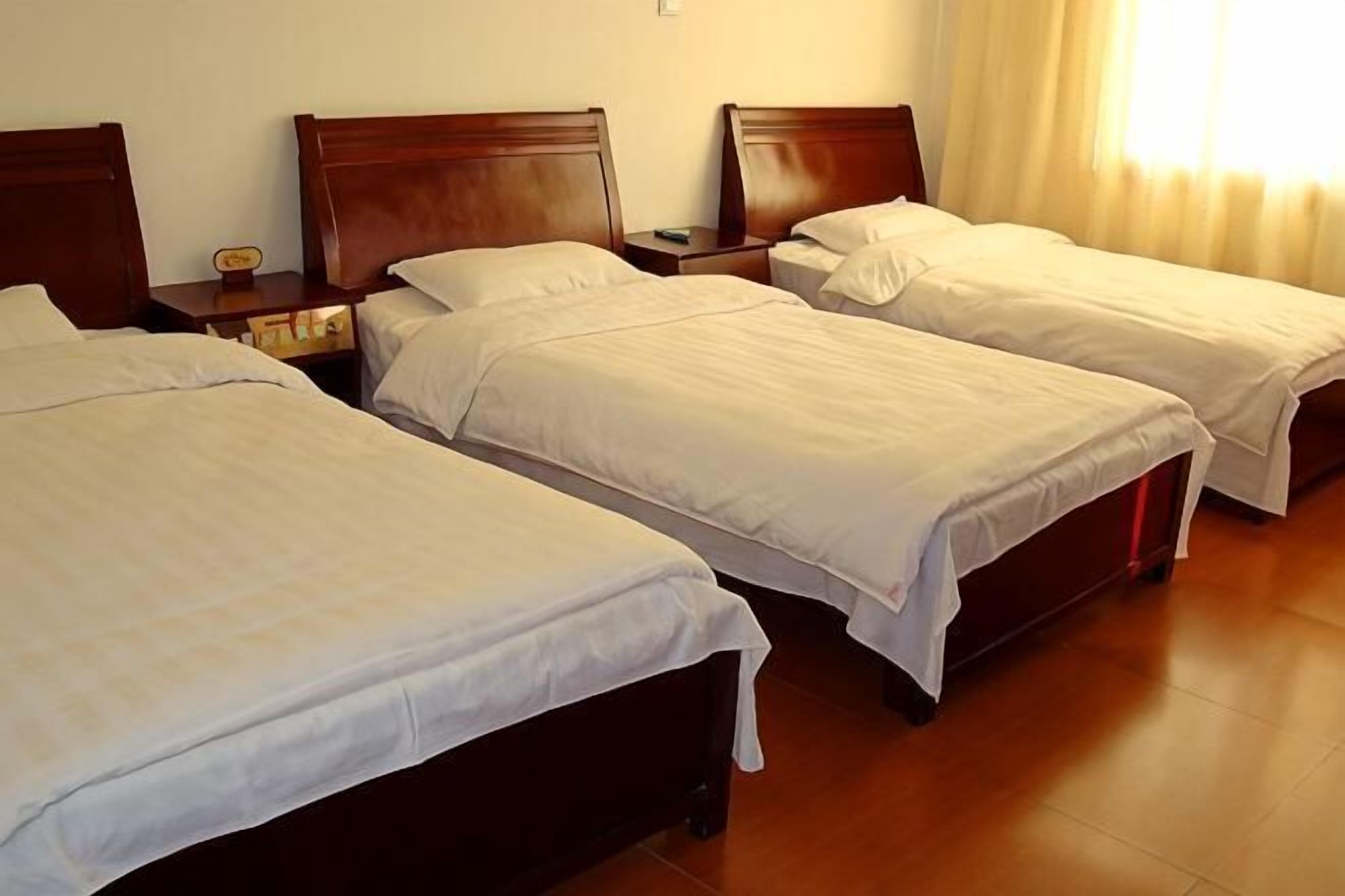 A hotel room with three single beds, wooden headboards, a nightstand with a lamp, and a window with curtains.