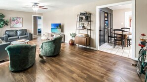 A spacious living room with hardwood flooring, a grey sofa, two green armchairs, a coffee table, and a TV displaying a beach scene.