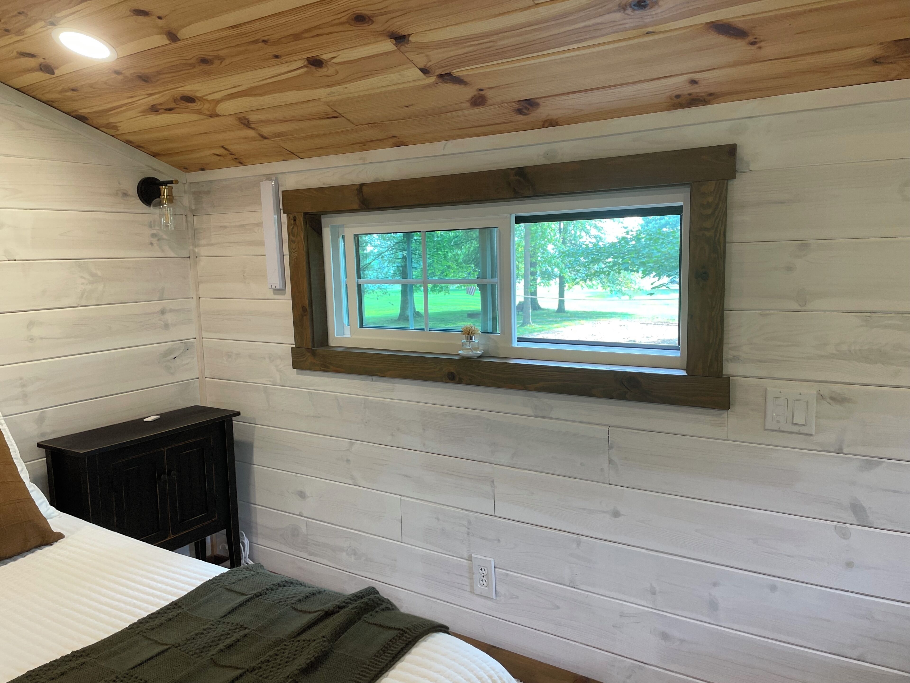 A bedroom with a wooden ceiling, a window with a view of trees, a bed with a green blanket, and a dark wooden cabinet.