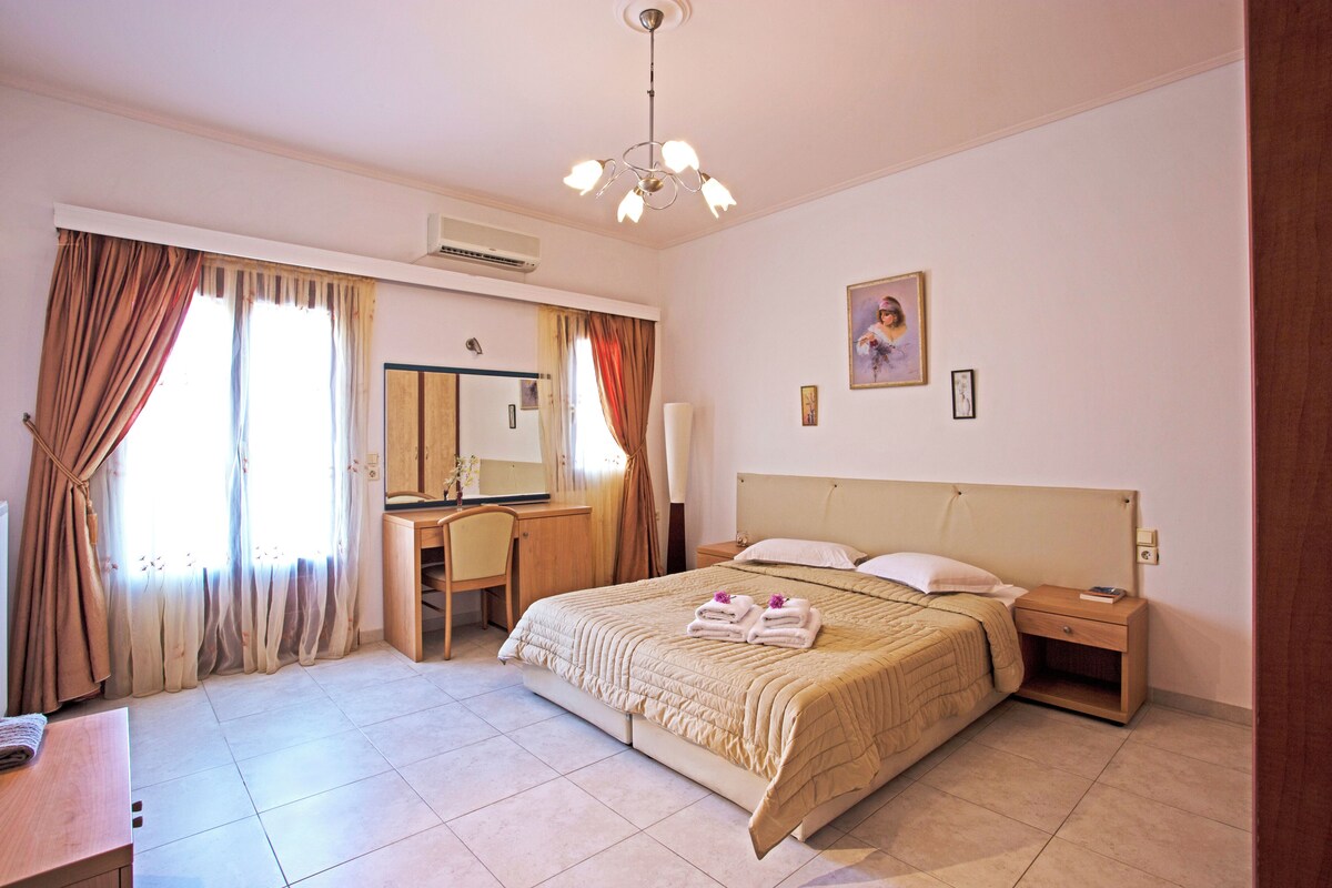 A hotel room with a bed, bedside table, nightstand, desk, chair, and a large window with curtains.