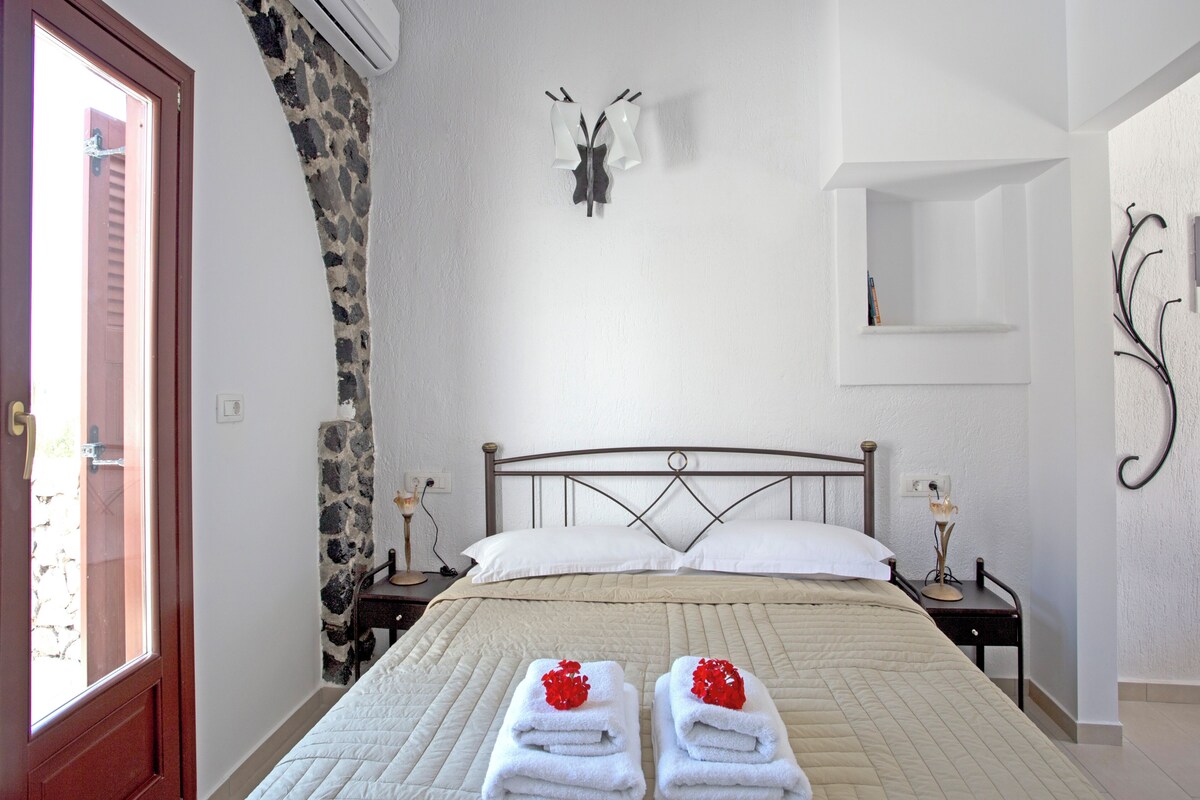 A neatly made bed with white linens and two towels with red flowers, a stone accent wall, a simple bedside table with a lamp, and a wall-mounted light fixture.