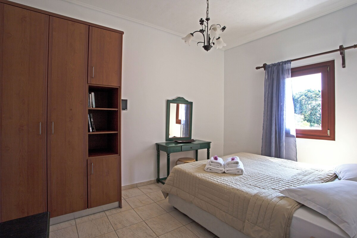 A bedroom with a bed, a wardrobe, a small table, a mirror, and a window with curtains.