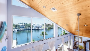 A room with large windows overlooking a body of water, wooden ceiling, and a view of a building across the water. - Sleeps 10 - Private house with 110’ of Deep Water Dockage, Pool, Hot Tub & more! (Marathon)