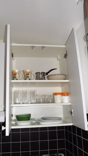 A kitchen cabinet with shelves containing various kitchenware, including mugs, a pot, bowls, and glasses.