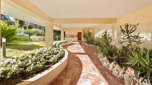 Un couloir dont le sol est incurvé, bordé de jardinières contenant différentes plantes et offrant une vue sur un jardin extérieur.