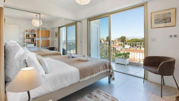Une chambre moderne avec un grand lit, un coin salon avec un fauteuil, un balcon avec vue et un lustre.