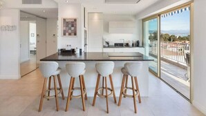 Une cuisine moderne dotée d’un îlot central et de tabourets de bar, de grandes fenêtres et d’une vue sur l’extérieur.
