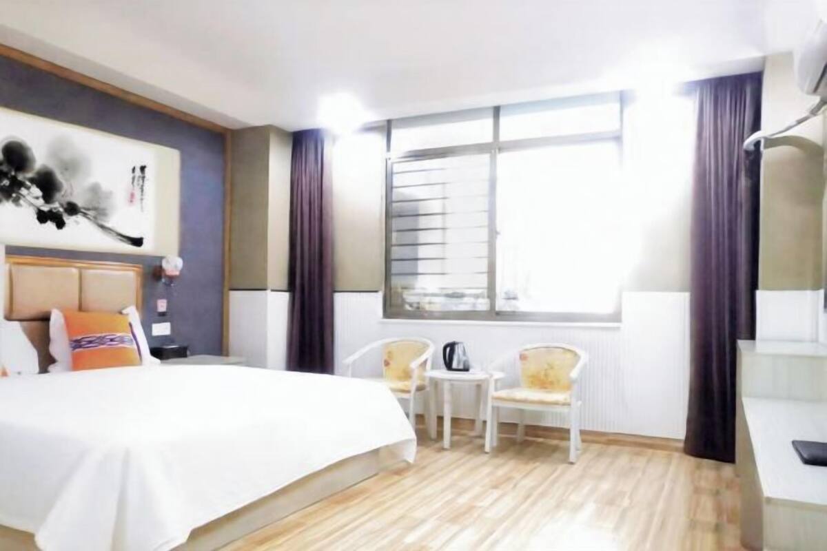 A hotel room with a bed, two chairs, a small table, a window with curtains, and a wall-mounted artwork.