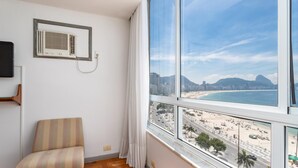 A room with a large window overlooking a beach, a chair, and a wall-mounted air conditioner. - View of Copacabana Beach A1103 (Rio de Janeiro)