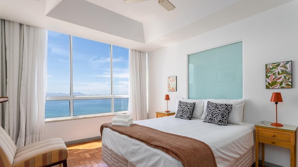 A hotel room with a large window, a bed with a brown blanket, two bedside tables with lamps, and a chair. - View of Copacabana Beach A1103 (Rio de Janeiro)