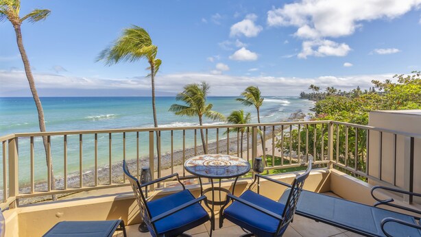 A balcony with a table and chairs, overlooking a beach with palm trees and the ocean. - The Paradise Penthouse (Lahaina)