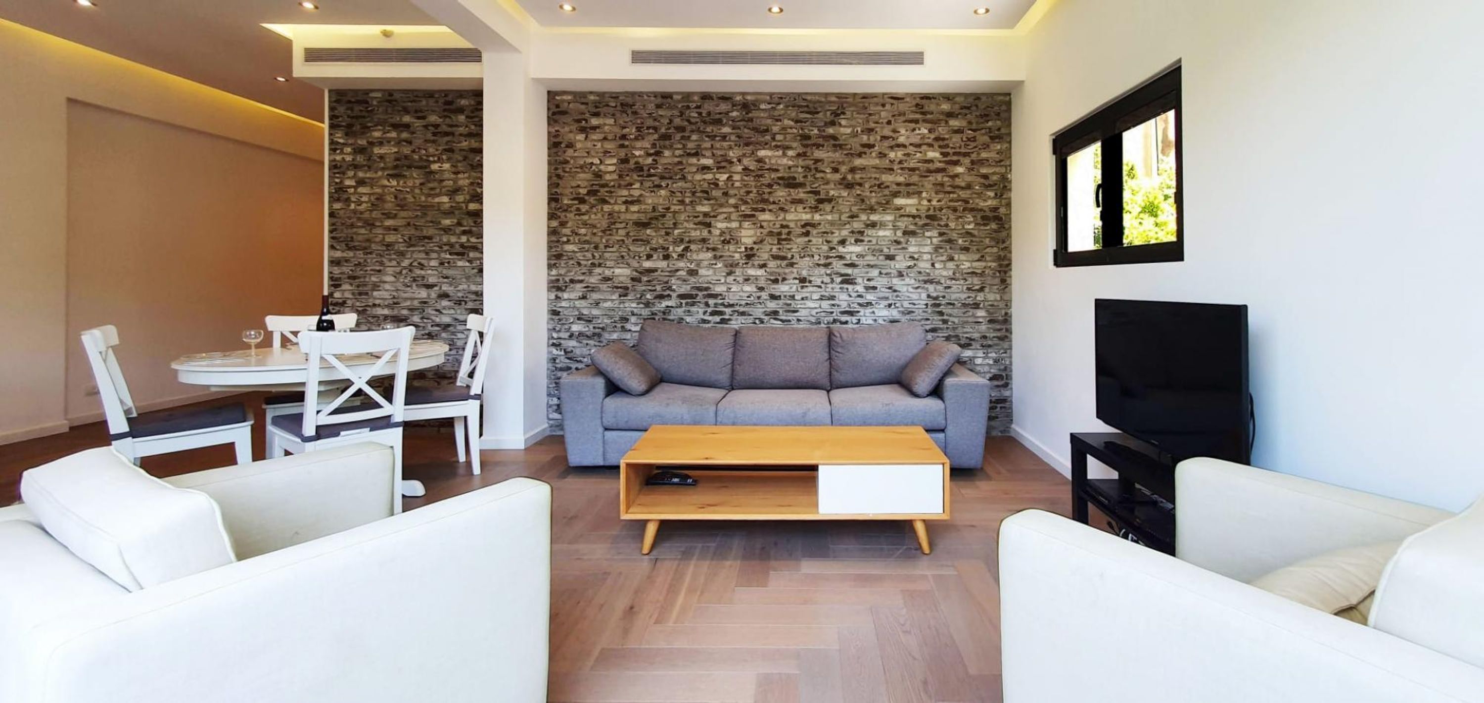 Een moderne woonkamer met een stenen muur, een grijze bank, witte stoelen, een houten salontafel en een televisie.