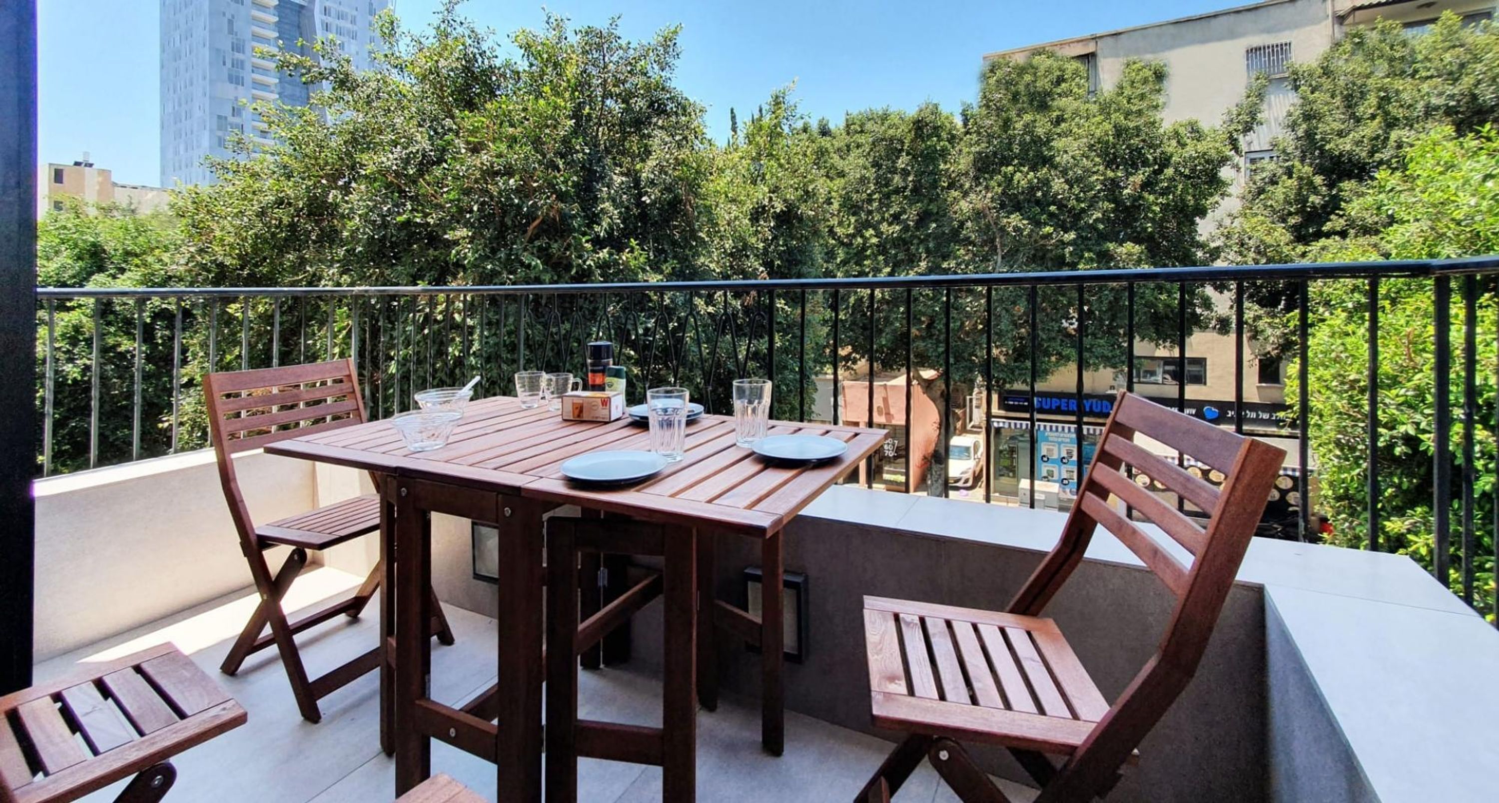 A balcony with a wooden table and chairs, surrounded by greenery and buildings.