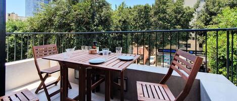 A balcony with a wooden table and chairs, surrounded by greenery and buildings.