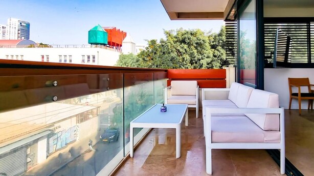 A modern balcony with a glass railing, white furniture, and a view of city buildings and trees. - Kalisher Ophrys in Tel Aviv-yafo (Tel Aviv)