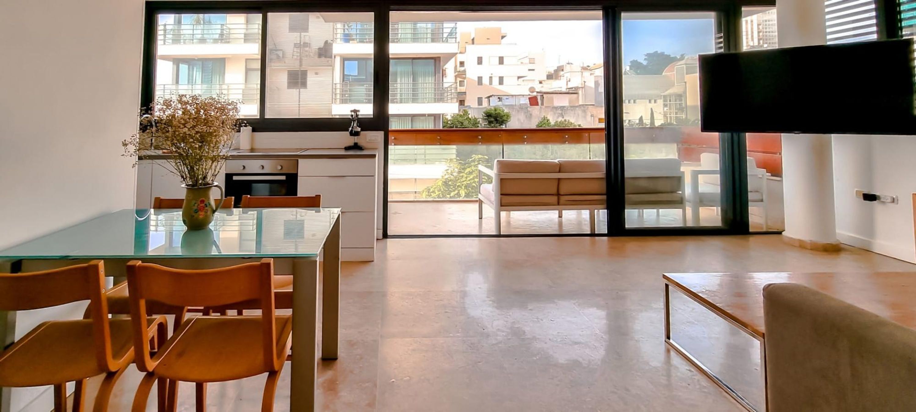 A modern kitchen with a glass dining table and chairs, a vase with dried flowers, and a view of the city through large windows.