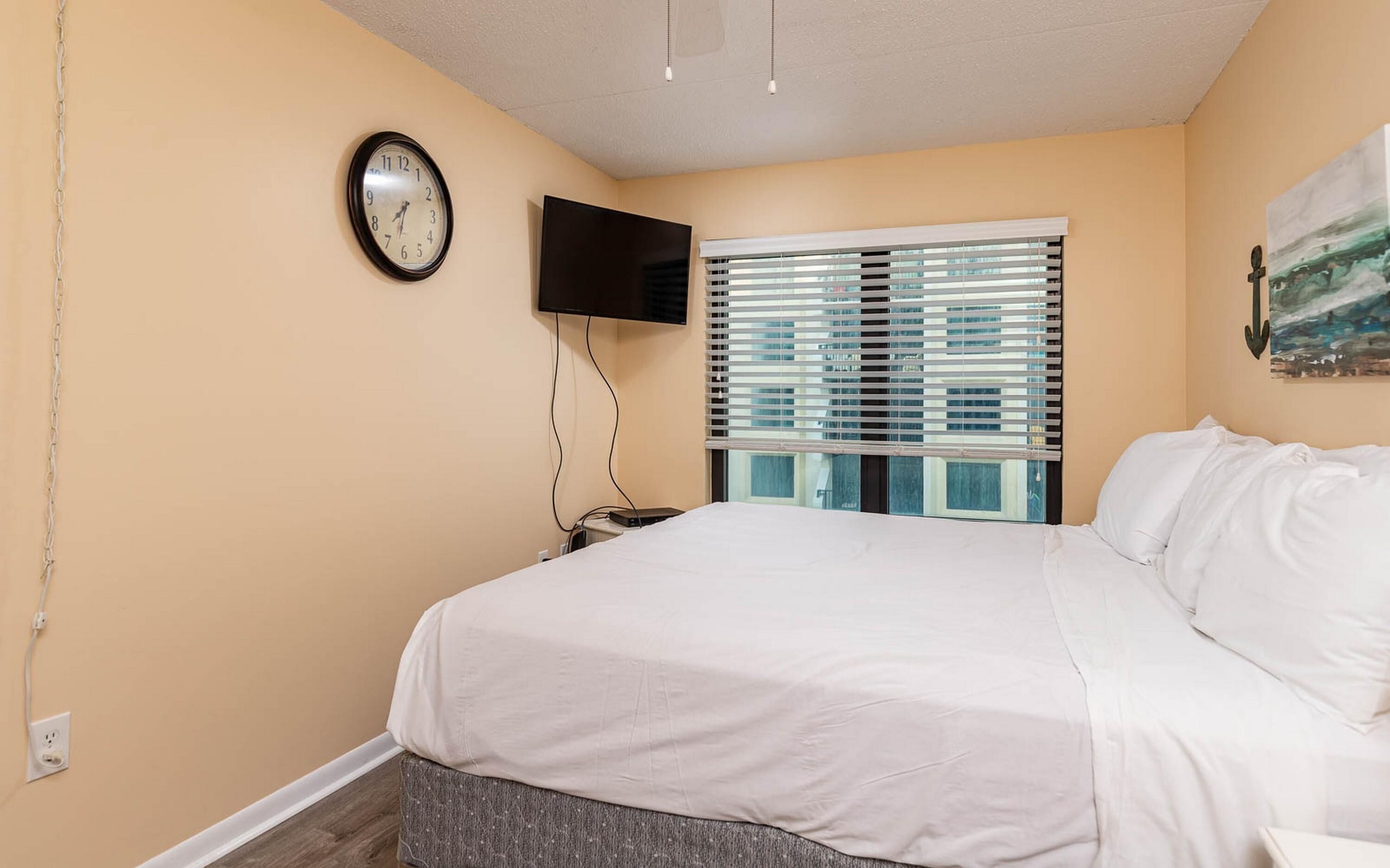 A bedroom with a bed, a wall-mounted TV, a window with blinds, and a clock.