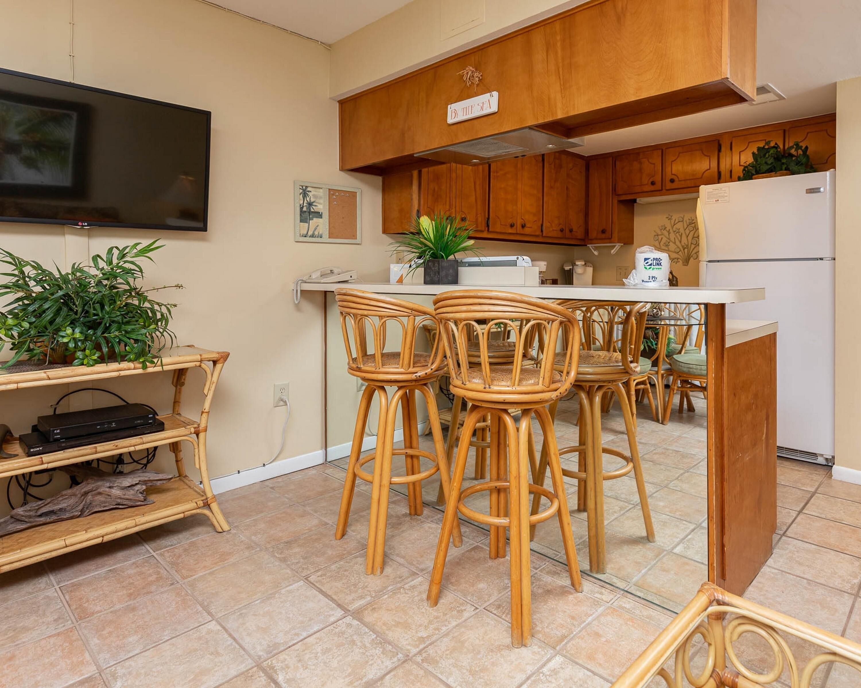 Una cocina con armarios de madera, un comedor con cuatro sillas, un televisor montado en la pared y un refrigerador.