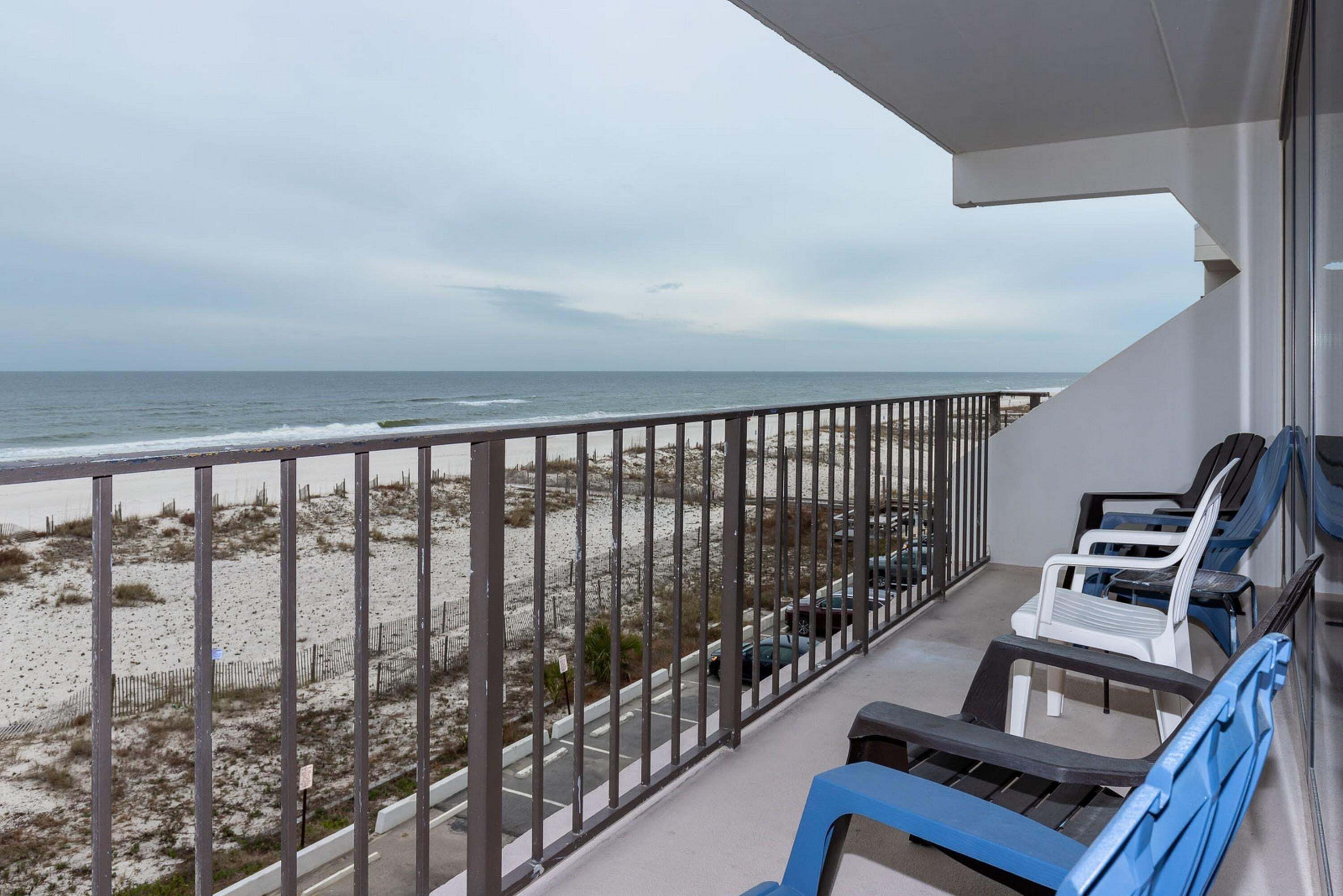A balcony with a view of the beach and ocean, a railing, and a few chairs.
