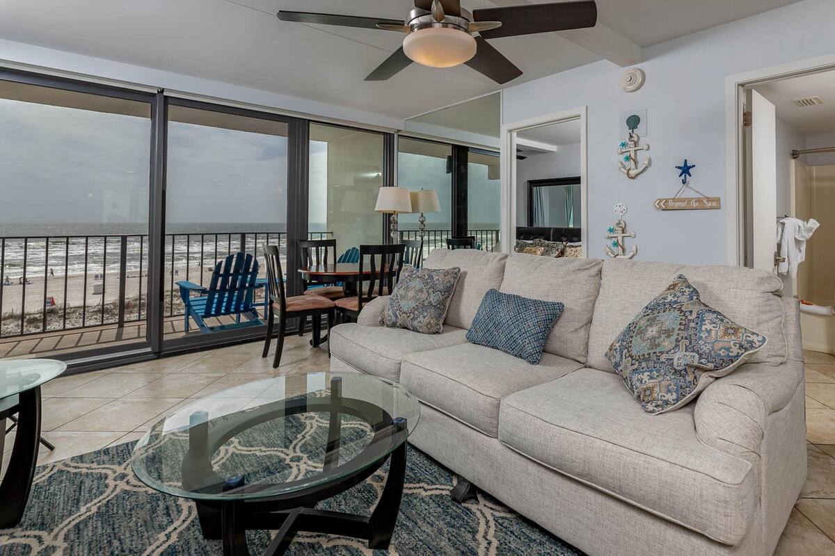 Ein Wohnzimmer mit einer Couch, einem Couchtisch und Sesseln. Es gibt einen Balkon mit Blick auf den Strand.