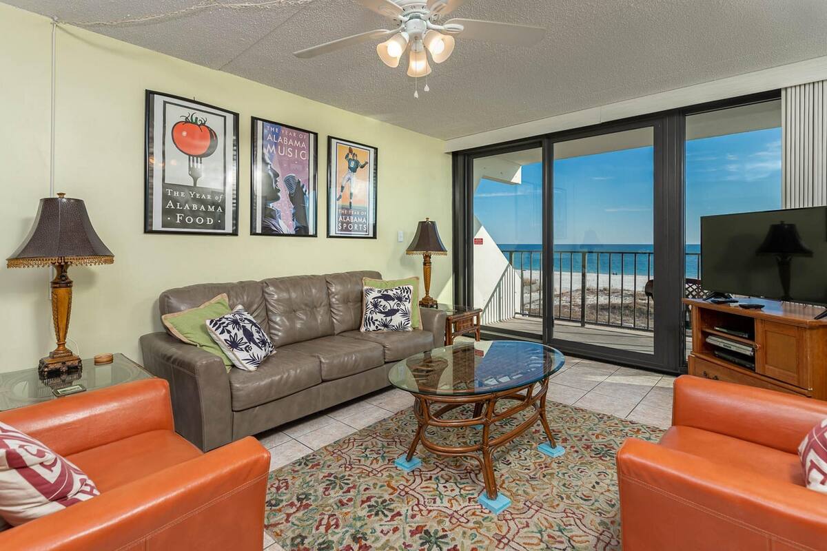 Ein Wohnzimmer mit einer Couch, zwei Sesseln, einem Glastisch, einem Fernseher und einem Balkon mit Blick auf den Strand.