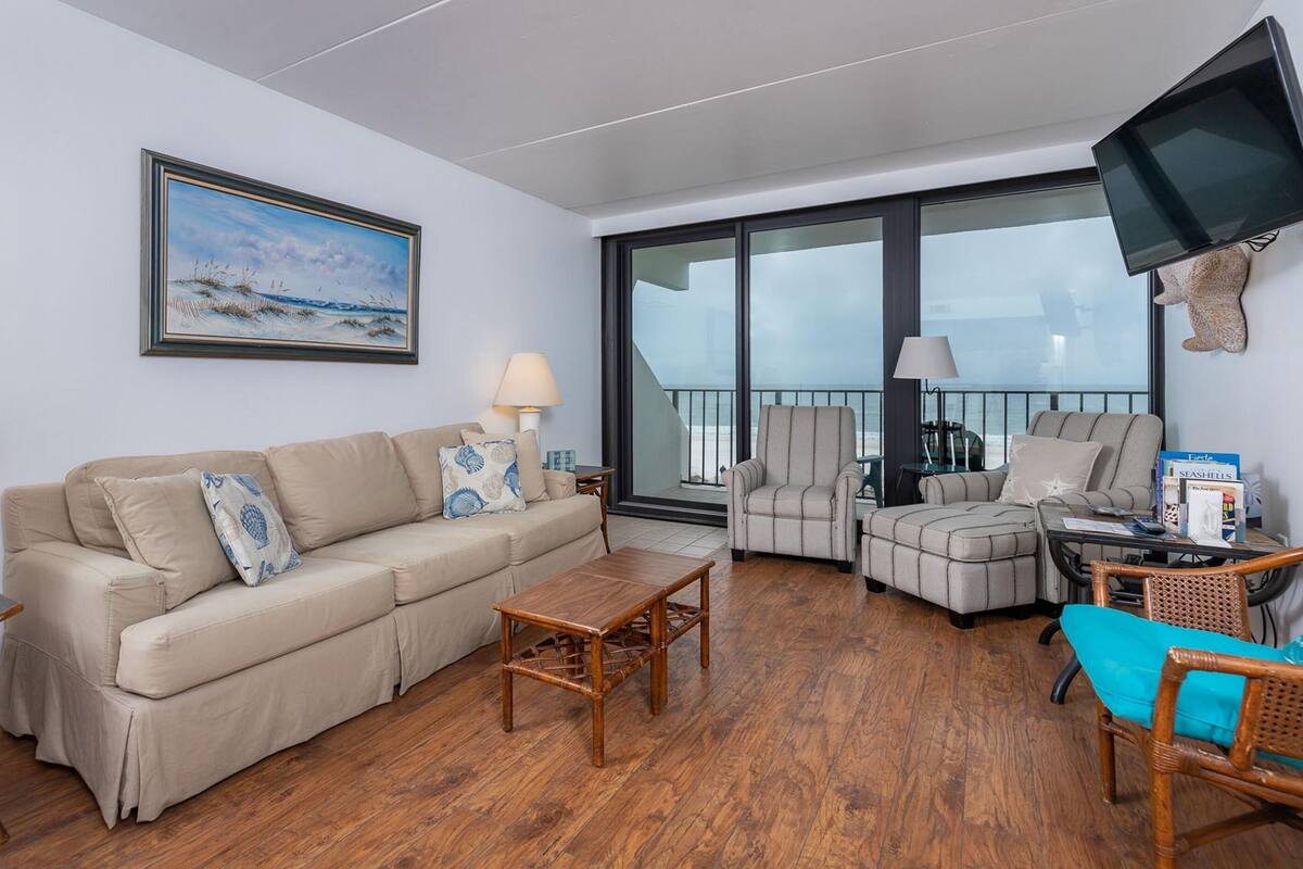 Ein Wohnzimmer mit einer Couch, Sesseln, einem Couchtisch und einem Fernseher. Ein großes Fenster bietet Blick aufs Meer.